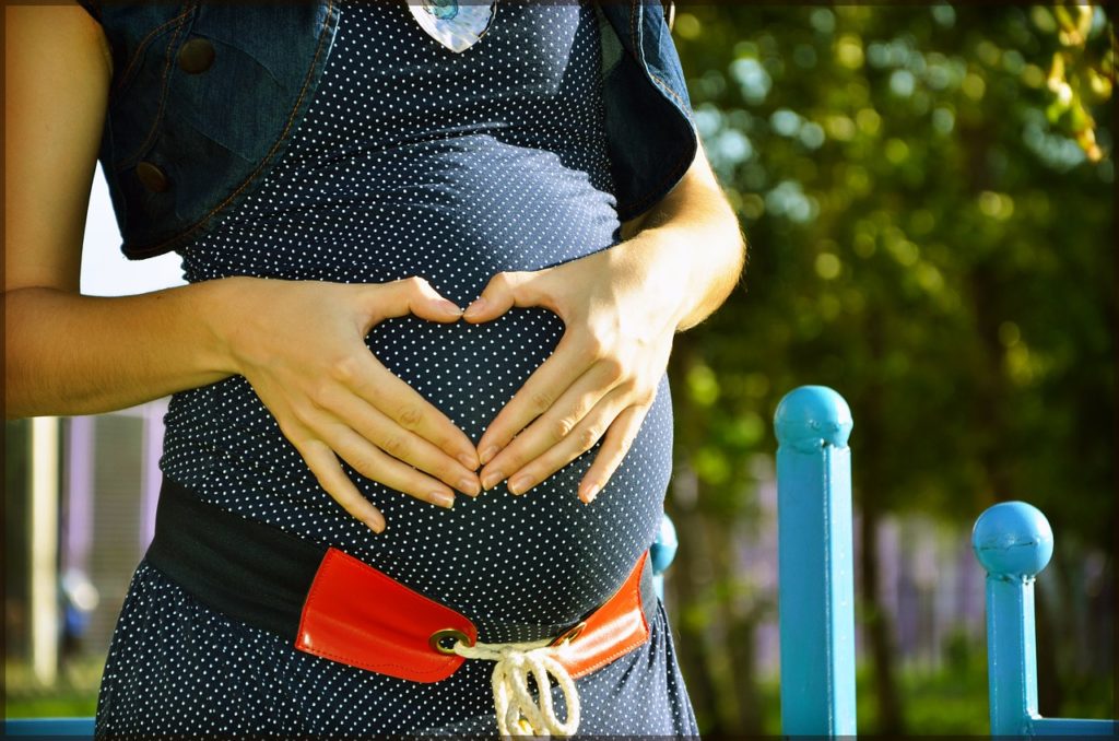 Schwangere fässt sich an den Bauch und formt ein Herz mit Händen