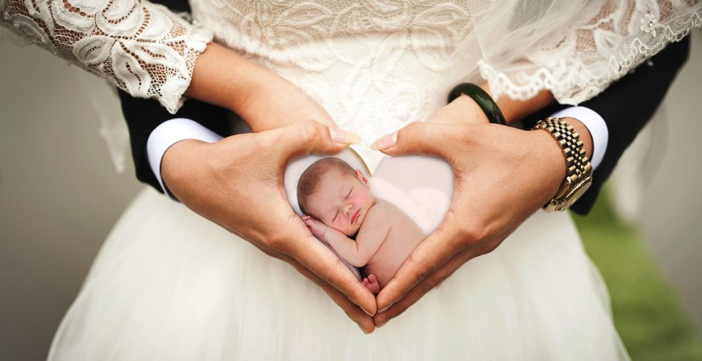 Braut und Bräutigam formen ein Herz mit den Händen am Bauch der Braut. Darin ein Babyfoto.